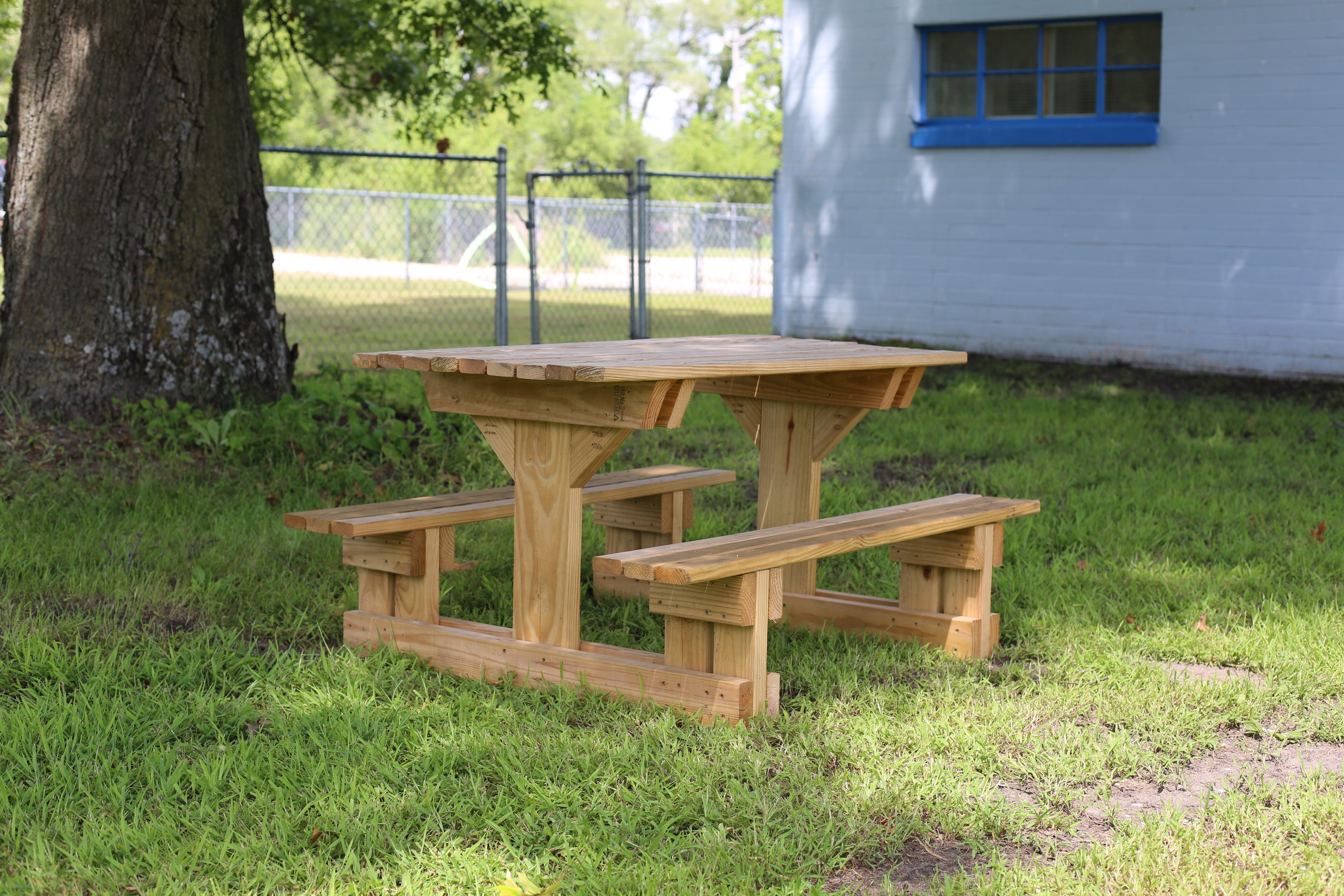 Wooden picnic table with benches on a grassy area near a building and tree.